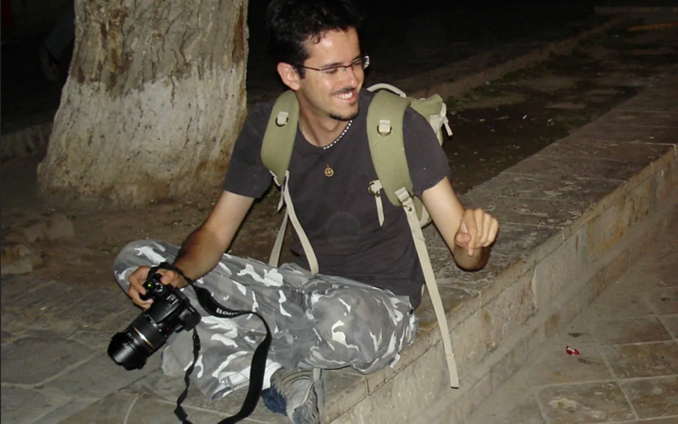 Giorgio sitting cross-legged on a stone ledge at night, a camera in his hand, laughing