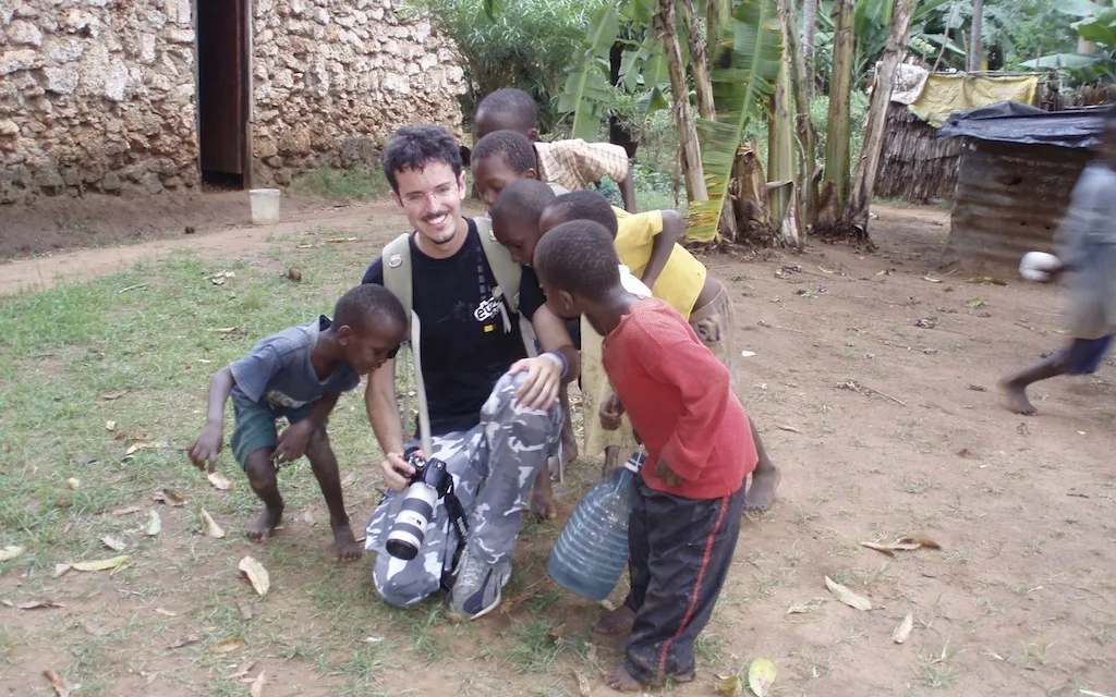 Giorgio crouching in a dirt yard in Kenya with a camera, children crowding round to see the screen